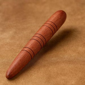 May include: A smooth, wooden massage tool with a tapered end and a series of carved rings. The tool is a warm reddish-brown color and appears to be made of a hardwood. The background is a textured, tan surface.