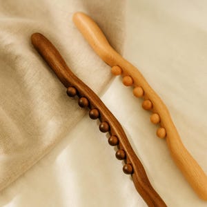 Peut inclure: Deux outils de massage en bois. L'un est brun foncé, l'autre est beige clair. Chacun a une poignée incurvée avec de petites boules en bois rondes. Ces outils sont conçus pour le massage musculaire et la relaxation. Mesure environ 30 cm de long.