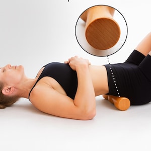 Pode incluir: Uma pessoa est&aacute; deitada de costas com uma ferramenta de massagem de madeira colocada sob a regi&atilde;o lombar. A ferramenta &eacute; cil&iacute;ndrica e feita de madeira clara. O texto "Rubber Grips for Traction" &eacute; vis&iacute;vel.
