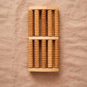 Peut inclure: Un masseur de pieds en bois avec plusieurs rouleaux à rainures. Le masseur est en bois de couleur claire et de forme rectangulaire. Les rouleaux sont disposés en rangées verticales et sont conçus pour masser les pieds. Le fond est un tissu beige neutre.