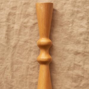 Puede incluir: Un candelero de madera clara con una forma de reloj de arena única. El candelero presenta una base y una parte superior ensanchadas, con una sección redondeada y bulbosa en el medio. La veta de la madera es visible, agregando textura al diseño.