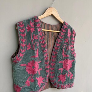 K&ouml;nnte beinhalten: Eine gr&uuml;ne Samtweste mit rosa Blumenstickerei. Die &auml;rmellose Weste hat ein gestreiftes Futter. Die Stickerei zeigt gro&szlig;e Blumen und Blattmuster und schafft ein dekoratives Design. Sie h&auml;ngt an einem Holzb&uuml;gel.
