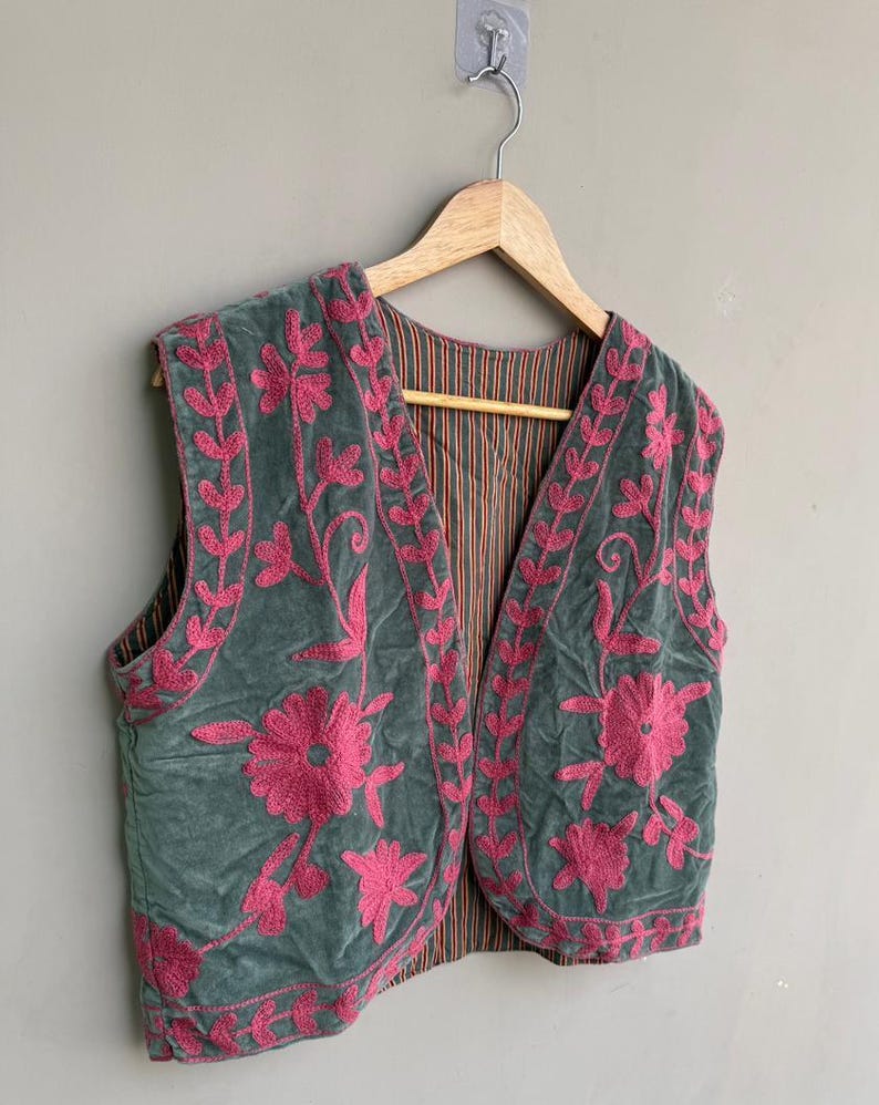 K&ouml;nnte beinhalten: Eine gr&uuml;ne Samtweste mit rosa Blumenstickerei. Die &auml;rmellose Weste hat ein gestreiftes Futter. Die Stickerei zeigt gro&szlig;e Blumen und Blattmuster und schafft ein dekoratives Design. Sie h&auml;ngt an einem Holzb&uuml;gel.