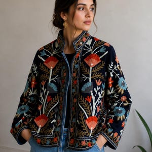 Pu&ograve; includere: Una giacca blu navy scuro con colletto alto e maniche lunghe, decorata con intricati ricami floreali nei toni dell'arancione, azzurro e bianco. La giacca ha una linea dritta ed &egrave; indossata sopra un top nero e un paio di jeans blu.