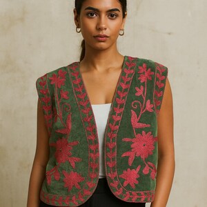 K&ouml;nnte beinhalten: Eine &auml;rmellose gr&uuml;ne Weste mit roter Blumenstickerei. Die Weste hat einen V-Ausschnitt und wird &uuml;ber einem wei&szlig;en Oberteil und einer schwarzen Hose getragen. Die Stickerei zeigt Blumen- und Bl&auml;ttermuster und verleiht dem Kleidungsst&uuml;ck eine dekorative Note.