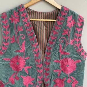 K&ouml;nnte beinhalten: Eine tealfarbene Samtweste mit leuchtend pinkfarbener Blumenstickerei. Die Weste hat ein gestreiftes Futter in Rot-, Braun- und Goldt&ouml;nen. Die Stickerei zeigt gro&szlig;e Blumen und zarte Bl&auml;ttermuster.