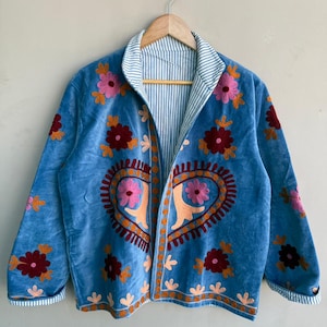May include: A light blue jacket with floral embroidery. The jacket features a heart-shaped design on the front and is adorned with pink, orange, and maroon flowers. The interior lining has a blue and white striped pattern. The jacket is hanging on a wooden hanger.