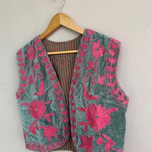 K&ouml;nnte beinhalten: Eine tealfarbene Samtweste mit leuchtend rosa Blumenstickerei. Die &auml;rmellose Weste h&auml;ngt an einem Holzb&uuml;gel. Das Innenfutter hat ein gestreiftes Muster in Orange- und Braunt&ouml;nen.