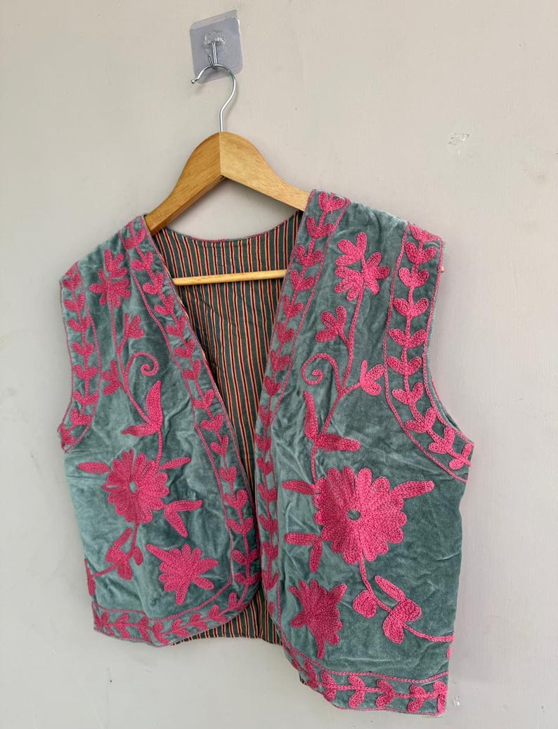 K&ouml;nnte beinhalten: Eine tealfarbene Samtweste mit leuchtend rosa Blumenstickerei. Die &auml;rmellose Weste h&auml;ngt an einem Holzb&uuml;gel. Das Innenfutter hat ein gestreiftes Muster in Orange- und Braunt&ouml;nen.