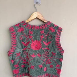 K&ouml;nnte beinhalten: Eine &auml;rmellose Weste in einem ged&auml;mpften Gr&uuml;nton, verziert mit leuchtend rosa Blumenstickereien. Die Stickerei zeigt eine zentrale Blume und Ranken, mit einem Rand aus floralen Designs. Die Weste h&auml;ngt an einem Holzb&uuml;gel.