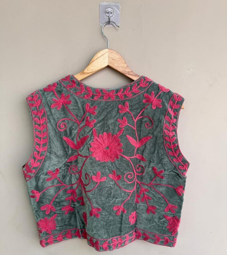 K&ouml;nnte beinhalten: Eine &auml;rmellose Weste in einem ged&auml;mpften Gr&uuml;nton, verziert mit leuchtend rosa Blumenstickereien. Die Stickerei zeigt eine zentrale Blume und Ranken, mit einem Rand aus floralen Designs. Die Weste h&auml;ngt an einem Holzb&uuml;gel.