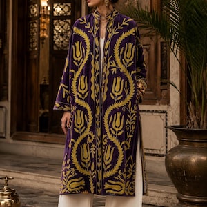 Pode incluir: Um casaco de veludo roxo profundo de manga comprida com um intrincado bordado floral amarelo brilhante num padrão vertical. O casaco tem gola alta, frente aberta e é usado sobre calças brancas lisas de perna larga.