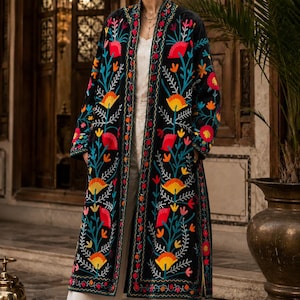 May include: A long black open-front coat featuring vibrant multi-colored floral embroidery in shades of red, yellow, and teal. The garment has long sleeves, a straight hem, and is worn over a white top and white wide-leg trousers with brown sandals.