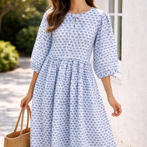 Puede incluir: Vestido estampado en azul claro y blanco, con cuello redondo y mangas tres cuartos. El vestido tiene cintura fruncida y falda a la rodilla. Se sostiene en la mano un bolso tote de paja tejida.
