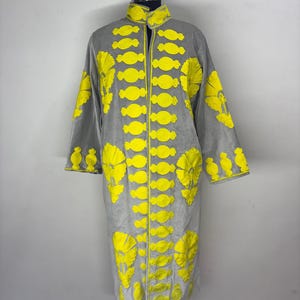 Può includere: Un cappotto grigio con dettagli ricamati in giallo brillante. Il cappotto presenta un collo alto e maniche lunghe. Il ricamo giallo include forme floreali e ovali, creando un contrasto sorprendente con il tessuto grigio. Il cappotto è esposto su un manichino.