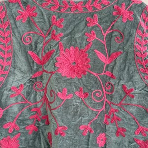 K&ouml;nnte beinhalten: Ein graues Samt-Oberteil mit leuchtend rosa Blumenstickerei. Die Stickerei zeigt eine gro&szlig;e zentrale Blume, zarte Ranken und Bl&auml;ttermuster. Das Oberteil wird auf einem Holzb&uuml;gel pr&auml;sentiert.