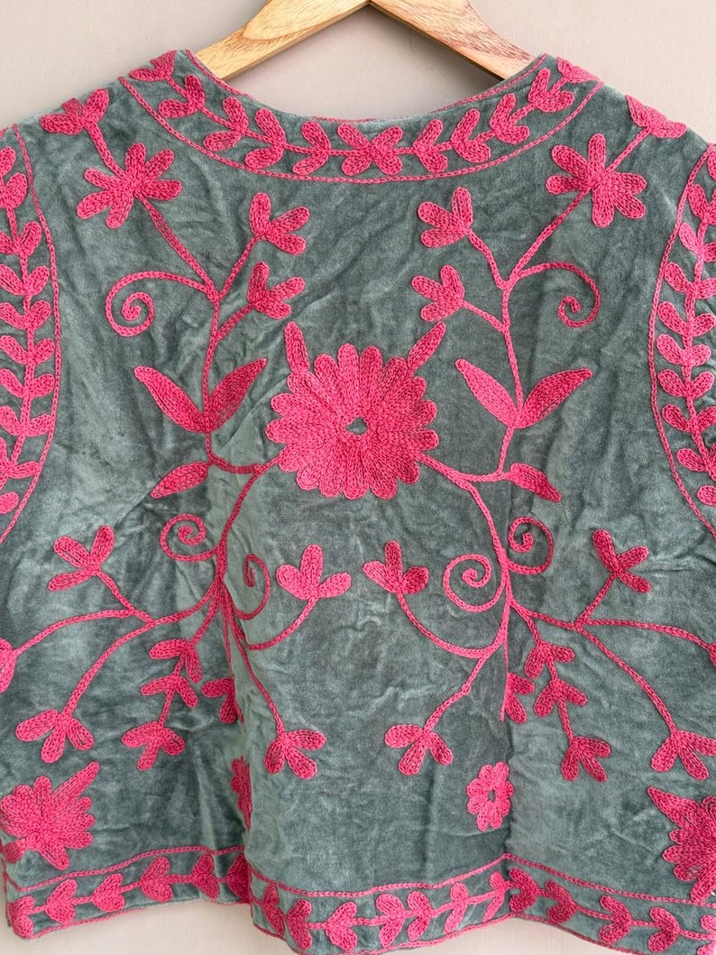 K&ouml;nnte beinhalten: Ein graues Samt-Oberteil mit leuchtend rosa Blumenstickerei. Die Stickerei zeigt eine gro&szlig;e zentrale Blume, zarte Ranken und Bl&auml;ttermuster. Das Oberteil wird auf einem Holzb&uuml;gel pr&auml;sentiert.