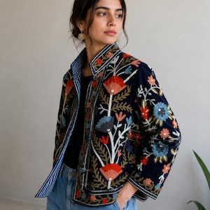 Pu&ograve; includere: Una giacca blu navy con vivaci ricami floreali nei toni del rosso, arancione e azzurro. La giacca presenta un colletto alto, maniche lunghe e una fodera a righe bianche e blu navy visibile lungo l'apertura frontale.