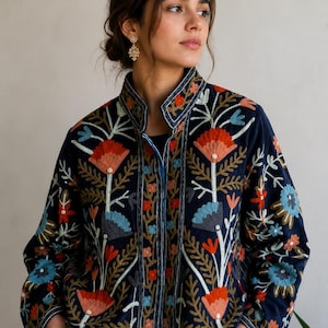 Pu&ograve; includere: Una giacca blu navy scuro caratterizzata da un intricato motivo floreale ricamato multicolore. Il design include grandi fiori arancioni e azzurri con steli e foglie verdi. La giacca presenta un colletto alto e maniche lunghe con ricami.
