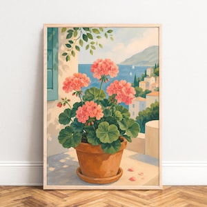 Op de afbeelding: Een ingelijste kunstdruk met een terracotta pot vol levendige roze geraniums. De afbeelding toont een kustgezicht met een blauwe zee, verre bergen en witte gebouwen onder een heldere lucht, alles gevat in een lichthouten lijst.