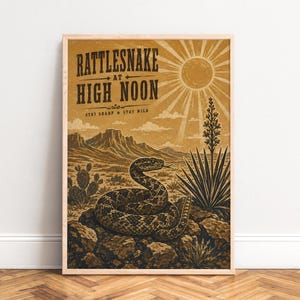 Puede incluir: Un póster enmarcado que muestra un paisaje desértico con una serpiente de cascabel enroscada sobre rocas, una planta de yuca alta y montañas lejanas bajo un sol brillante. El texto dice RATTLESNAKE AT HIGH NOON y STAY SHARP STAY WILD.