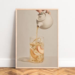 Op de afbeelding: Een ingelijste wandkunstprint met een beige theepot die gouden ijsthee in een geribbeld glas schenkt. Het glas bevat heldere ijsblokjes en twee verse schijfjes perzik. De compositie is geplaatst tegen een neutrale, effen beige achtergrond.