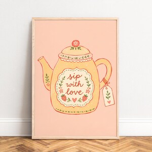 Op de afbeelding: Een rechthoekige wandkunstprint op een licht perzikkleurige achtergrond met een illustratie van een gele theepot. De theepot heeft een geschulpt label met de tekst 'sip with love', versierd met rode aardbeien, groene bladeren en een label.