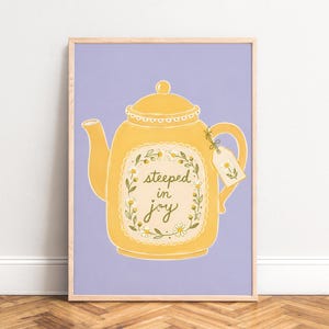 Puede incluir: Una lámina enmarcada con una tetera amarilla sobre un fondo morado claro. La tetera tiene un diseño de corona floral que rodea el texto "steeped in joy" y una pequeña etiqueta atada al asa con una ilustración de una margarita.