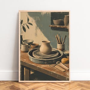 May include: A framed wall art print depicting a pottery studio scene with a clay vase on a wheel, various ceramic bowls, a mug, and wooden sculpting tools arranged on a rustic wooden workbench against a muted green and beige painted wall.