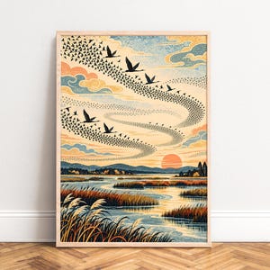 Op de afbeelding: Een ingelijste kunstdruk met een landschap met een kronkelende rivier, hoog gras op de voorgrond en verre bergen onder een zonsonderganghemel. Een grote zwerm vogels vliegt in een wervelende formatie door de lucht in tinten oranje, blauw en crème.