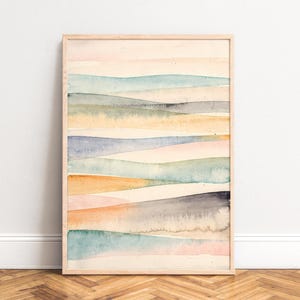 May include: Watercolor landscape art print in a light wood frame. The artwork features horizontal bands of color in shades of peach, blue, yellow, and gray. The abstract design evokes a sense of calm and natural beauty, suitable for home decor.