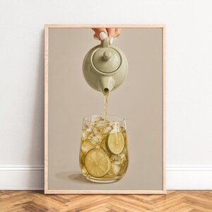 Peut inclure: Une affiche encadrée représentant une théière vert sauge versant un liquide doré dans un verre transparent rempli de glaçons et de deux rondelles de citron vert. La composition est placée sur un fond beige neutre et uni, créant un rendu épuré.