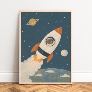 Könnte beinhalten: Ein gerahmter Kunstdruck mit einem braunen Bären in einer weiß-orangen Weltraumrakete, die durch einen dunkelblauen Sternenhimmel fliegt. Die Rakete hinterlässt eine weiße Rauchfahne über einem grün-blauen Planeten, mit einem Ringplaneten und Mond.