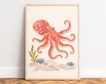 May include: A framed wall art print featuring a watercolor illustration of a coral-colored octopus with white spots. The octopus is positioned above a sandy seabed with small gray rocks and a sprig of green seaweed on a cream background.
