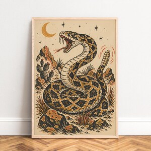 Op de afbeelding: Een ingelijste muurkunstprint met een gedetailleerde illustratie van een opgerolde ratelslang met een open bek en zichtbare giftanden. De slang bevindt zich in een woestijnlandschap met cactussen, rotsen, een maansikkel en sterren tegen een beige achtergrond.