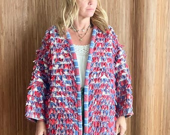May include: A long-sleeved jacket featuring a textured, fringed surface in a red, white, and blue pattern. The jacket has a blue and white striped trim along the front opening. It is worn over a white top and solid green wide-leg trousers.
