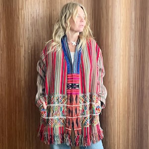 Puede incluir: Chaqueta a rayas colorida con dobladillo con flecos. La chaqueta presenta rayas verticales en rojo, rosa y beige, con un panel tejido detallado y un cuello azul. El dobladillo inferior tiene una colorida franja.