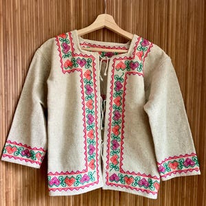 May include: A beige, open-front jacket with three-quarter sleeves. The jacket features vibrant embroidery in pink, orange, and green along the edges, neckline, and sleeves. It hangs on a wooden hanger against a wooden background.