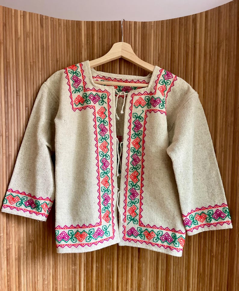 May include: A beige, open-front jacket with three-quarter sleeves. The jacket features vibrant embroidery in pink, orange, and green along the edges, neckline, and sleeves. It hangs on a wooden hanger against a wooden background.