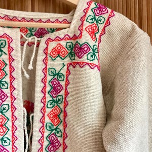 May include: Beige jacket with colorful floral embroidery in red, green, and pink. The jacket features a square neckline and a tie closure. The embroidery is detailed and covers the collar and edges of the jacket.