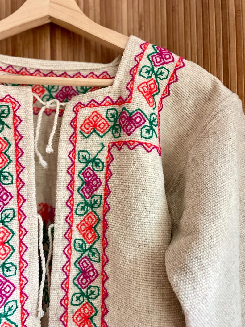 May include: Beige jacket with colorful floral embroidery in red, green, and pink. The jacket features a square neckline and a tie closure. The embroidery is detailed and covers the collar and edges of the jacket.