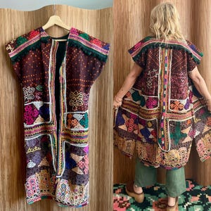 May include: A sleeveless, open-front long vest in dark brown fabric featuring intricate, multi-colored geometric embroidery, small mirror accents, and a dark green pom-pom trim along the shoulders. The garment displays a patchwork-style design.