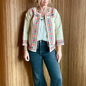 May include: Beige embroidered jacket with a square neckline and three-quarter sleeves. The jacket features colorful floral embroidery in pink, orange, and green along the edges. Worn with a white top, dark wash jeans, and platform shoes.