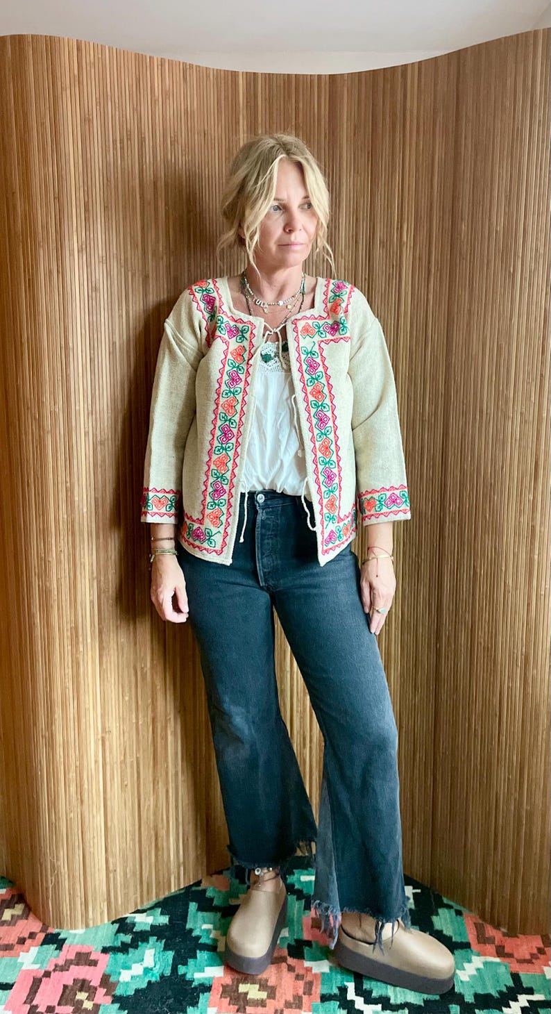 May include: Beige embroidered jacket with a square neckline and three-quarter sleeves. The jacket features colorful floral embroidery in pink, orange, and green along the edges. Worn with a white top, dark wash jeans, and platform shoes.