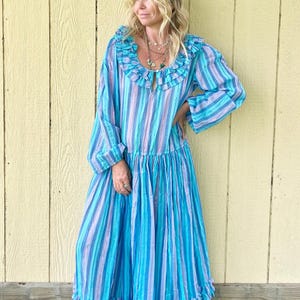 Puede incluir: Un vestido largo y fluido con rayas verticales en tonos azules y lavanda. El vestido presenta un cuello y un dobladillo con volantes, con mangas largas. La modelo lleva varios collares y zapatos marrones. El vestido está hecho de tela ligera.