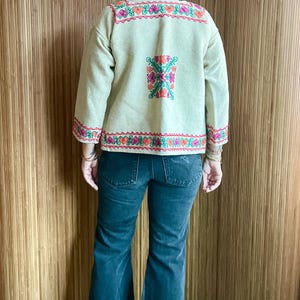 May include: Beige embroidered jacket with colorful floral and geometric patterns. The jacket features long sleeves and is paired with dark wash denim jeans. The embroidery includes pink, orange, and green details.