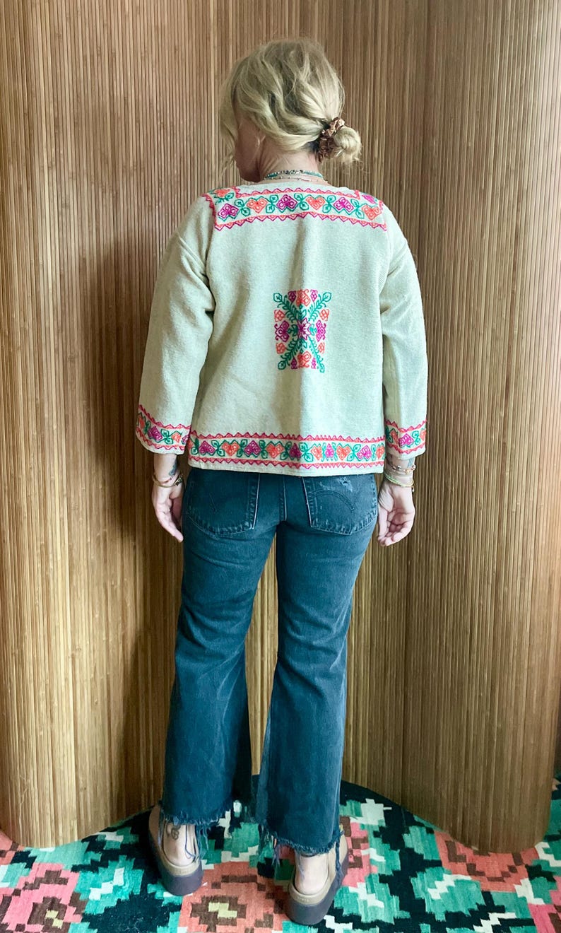May include: Beige embroidered jacket with colorful floral and geometric patterns. The jacket features long sleeves and is paired with dark wash denim jeans. The embroidery includes pink, orange, and green details.