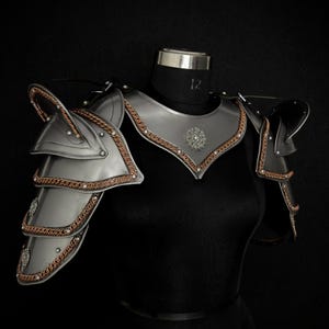 May include: A metallic silver shoulder and chest armor set featuring layered pauldrons with copper-colored chainmail trim. The armor includes a central chest plate adorned with a circular floral emblem and is displayed on a black mannequin torso.
