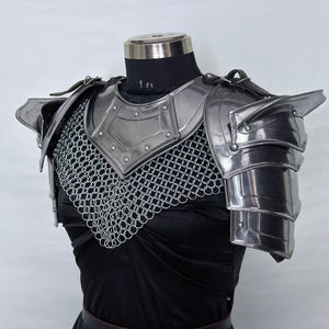 May include: Steel armor breastplate and shoulder guards with chainmail. The metal armor is silver and has a polished finish. The shoulder guards are layered and articulated. The armor is displayed on a mannequin with a black top and skirt. The logo "STEEL HERITAGE BATTLE ARMOR" is visible.