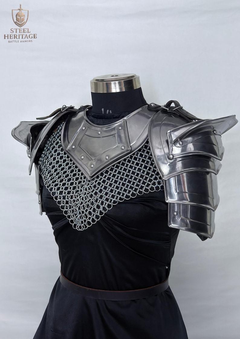 May include: Steel armor breastplate and shoulder guards with chainmail. The metal armor is silver and has a polished finish. The shoulder guards are layered and articulated. The armor is displayed on a mannequin with a black top and skirt. The logo "STEEL HERITAGE BATTLE ARMOR" is visible.